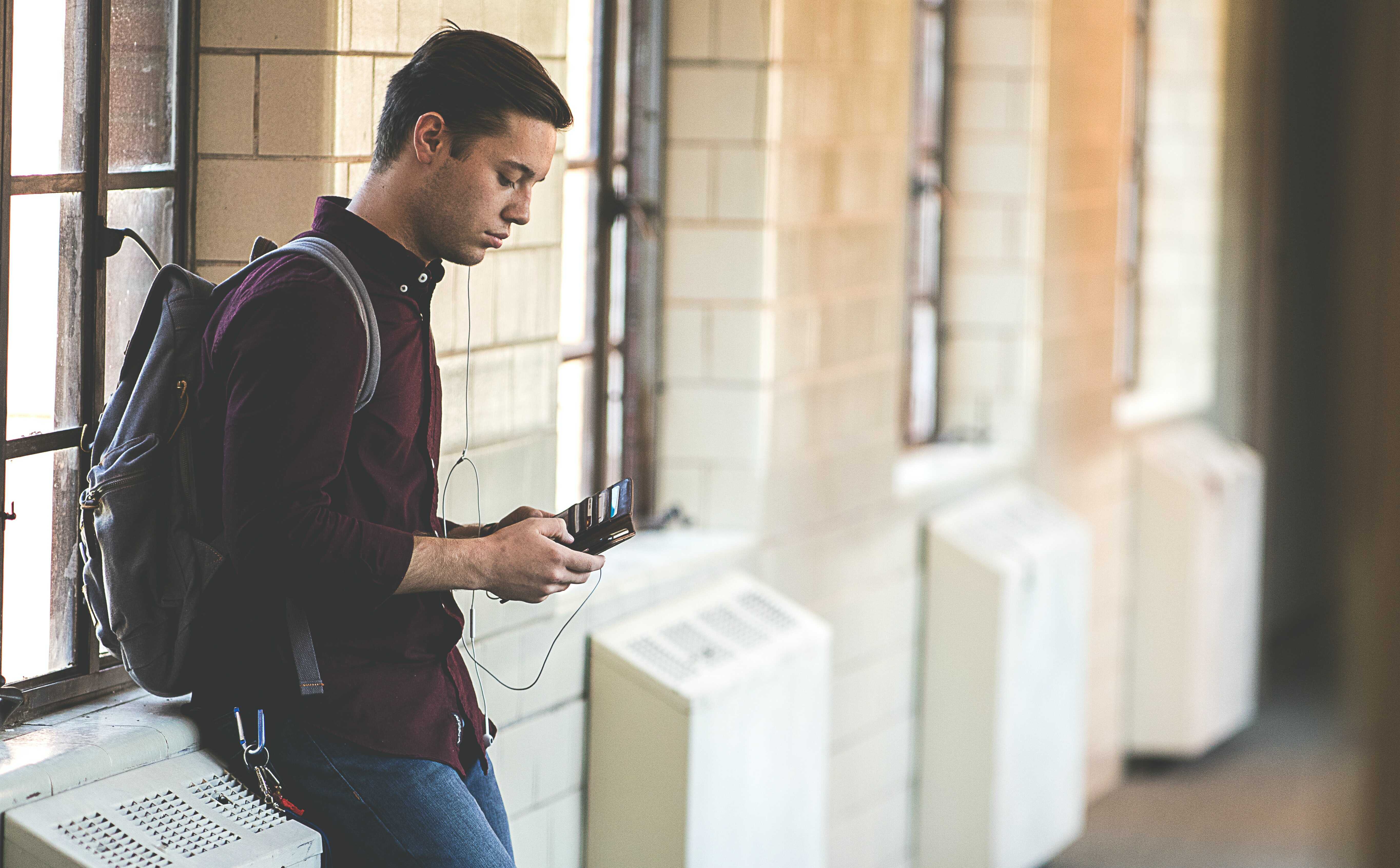 A young man with a backpack is on his phone in a hallway.